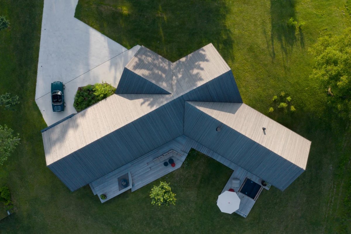 Aerial view of a multi-wing house with light gray, angular roof, set in a green lawn; driveway with a car and a wooden deck with an umbrella.