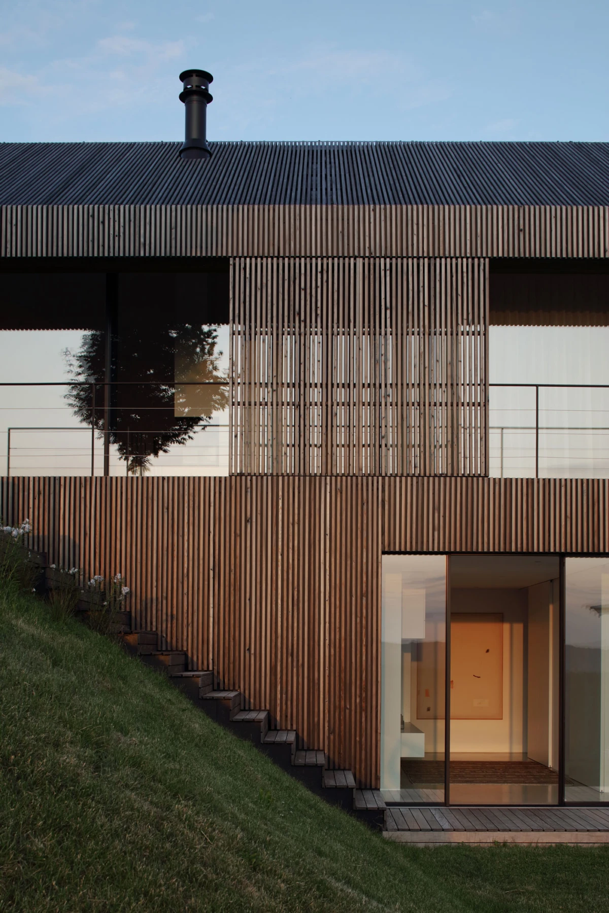 Front view of a modern wooden building with vertical slats and glass facade; hillside lawn with wooden stairs on the left and an illuminated entrance, rooftop chimney.