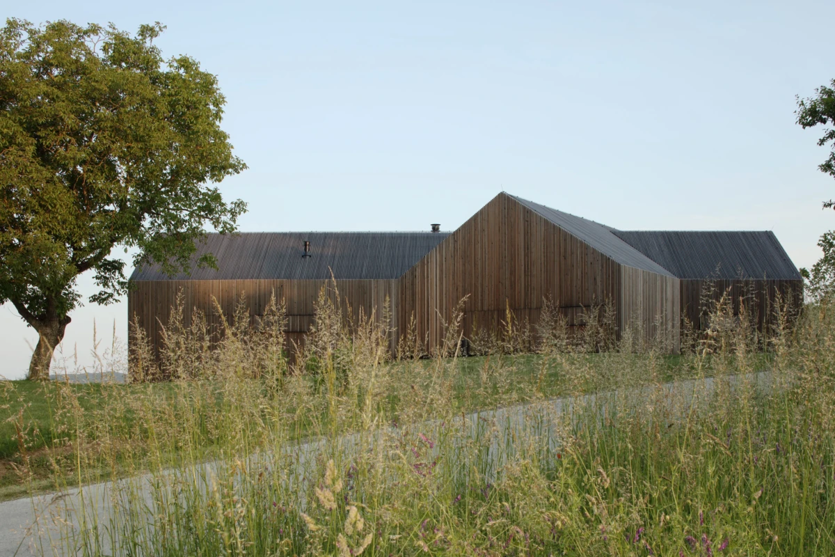 Wooden barn-like building sits in a grassy field beside a tree, with a gravel path in the foreground.