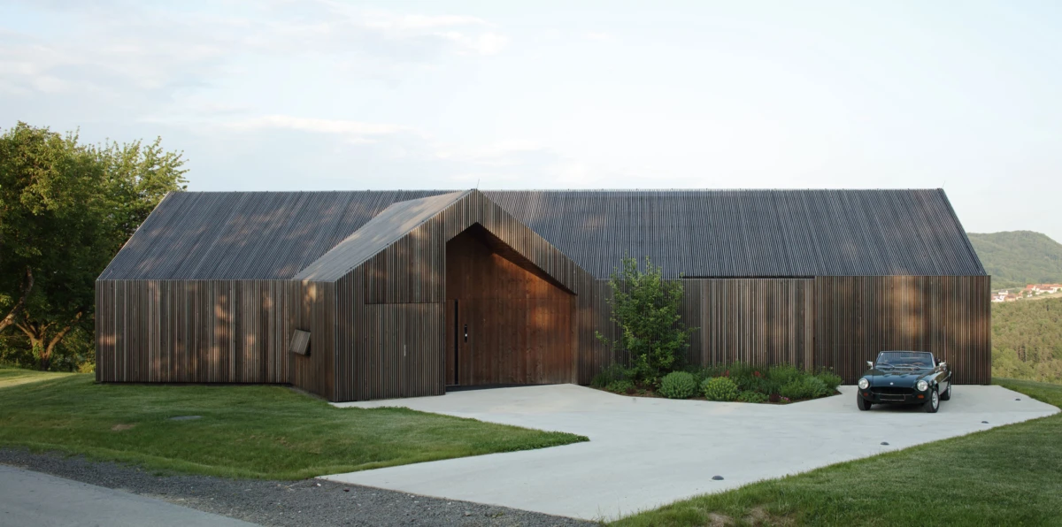 Modern dark-wood residence with a steep sloped roof and central entry; curved driveway, landscaped shrubs, and a black convertible parked outside.