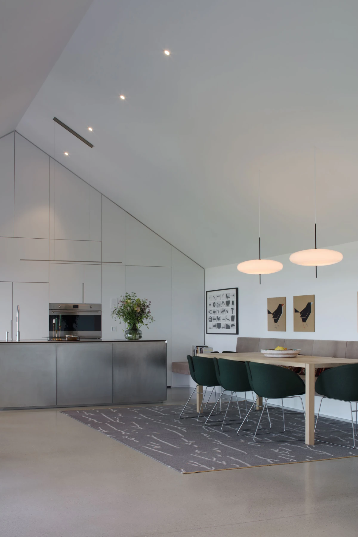 Modern kitchen and dining area with a stainless steel island, white cabinetry, and a wooden table with dark green chairs under pendant lights.