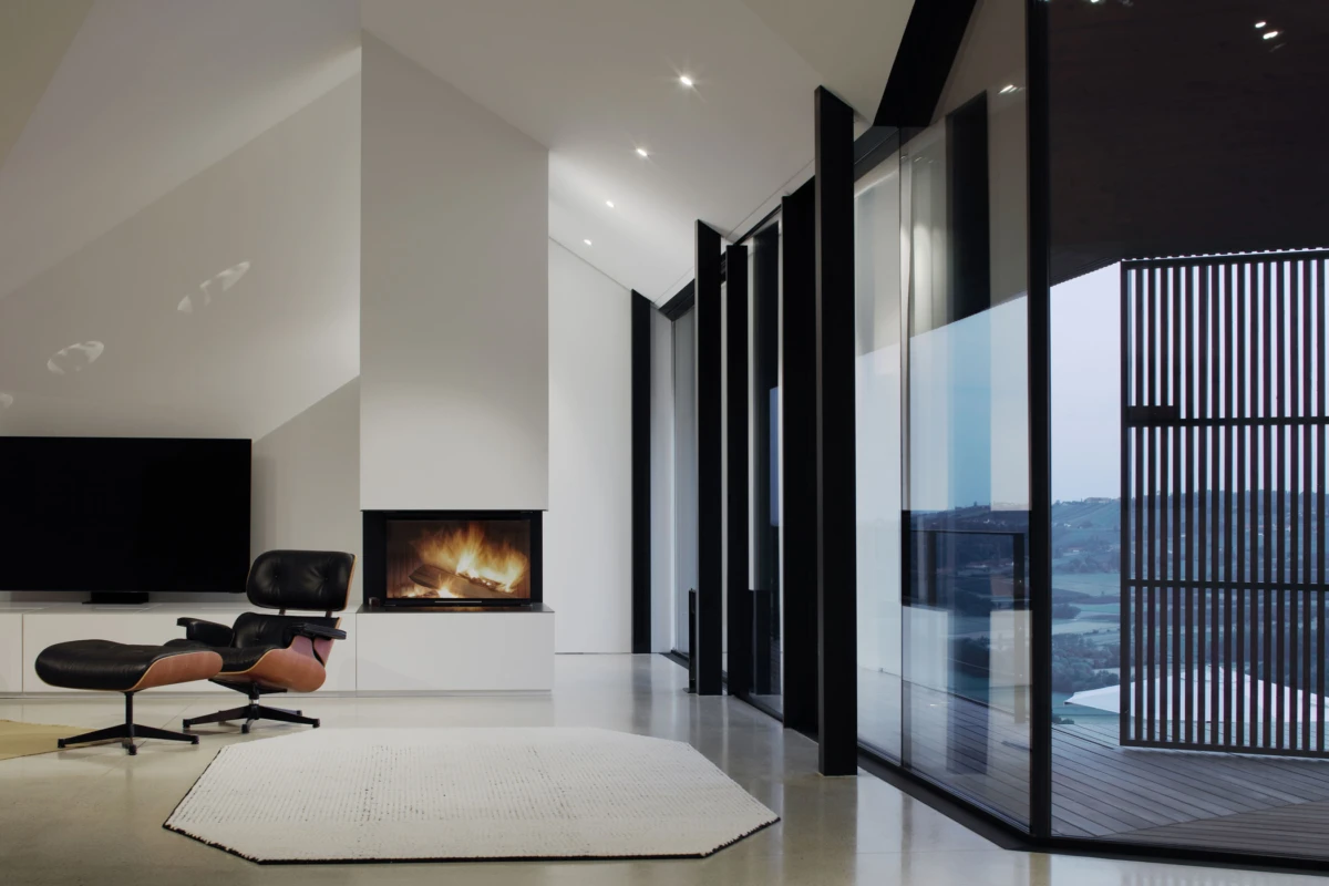 Minimalist living room with a built-in fireplace, Eames lounge chair and ottoman, and floor-to-ceiling glass walls framing a deck and distant hills.