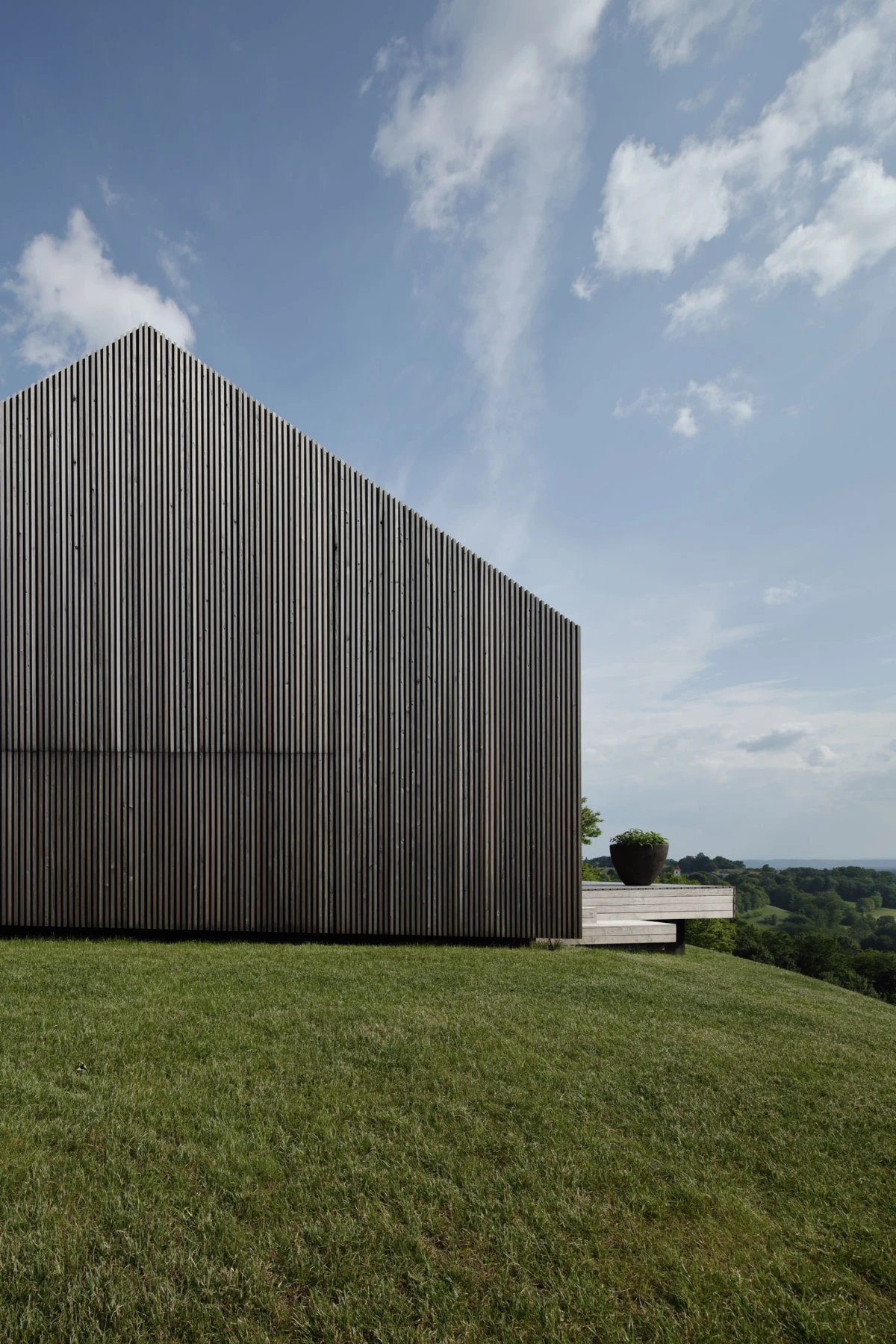 Wooden-clad building with vertical slats on a grassy hill, a deck with planters to the right, under a blue sky.
