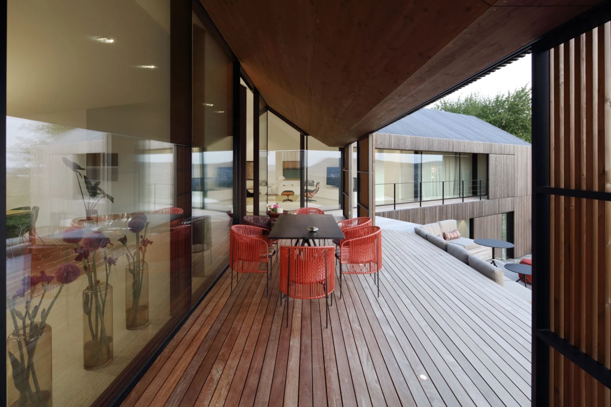 Red wicker chairs surround a dark rectangular table on a wooden balcony of a modern glass-walled house.