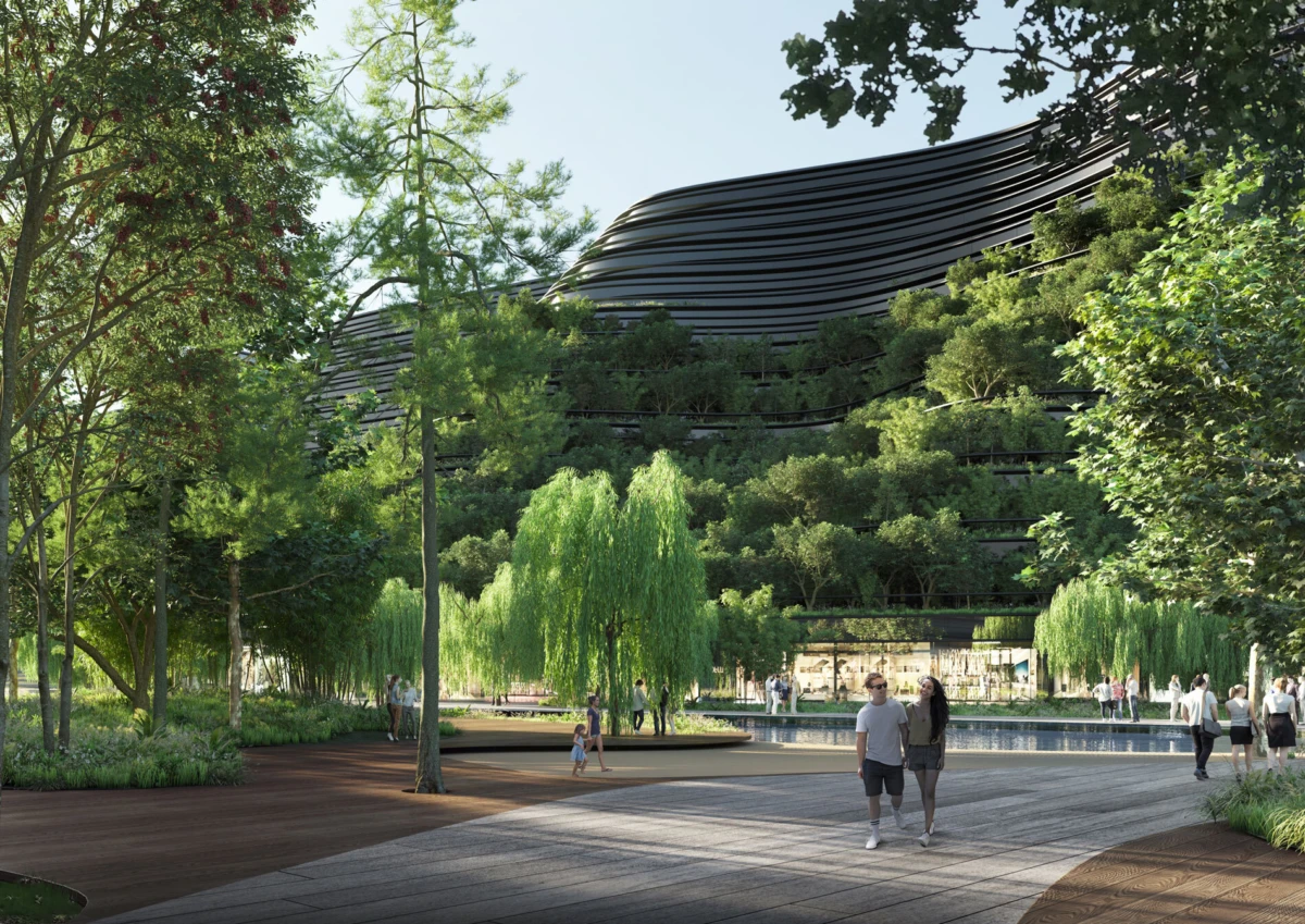 Curved black modern building rises behind a lush, tree-filled park; people stroll along a wooden boardwalk beside a shallow water feature.