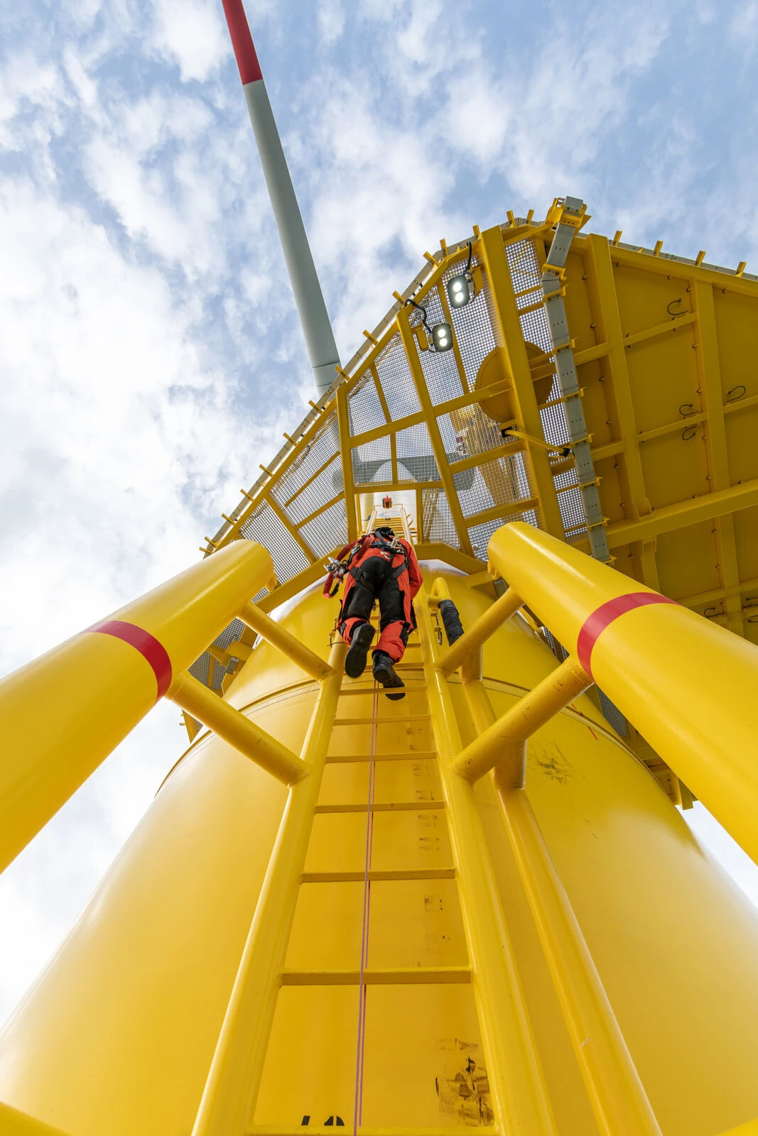 Worker in red protective gear climbs a yellow steel ladder on an industrial structure with a platform and blue sky.