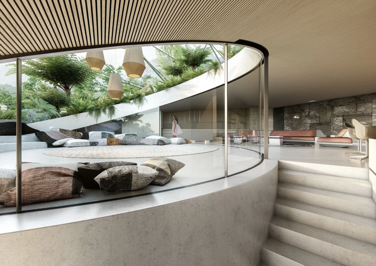 Person standing in a glass-walled circular lounge within a modern atrium, surrounded by plush cushions and hanging lamps.