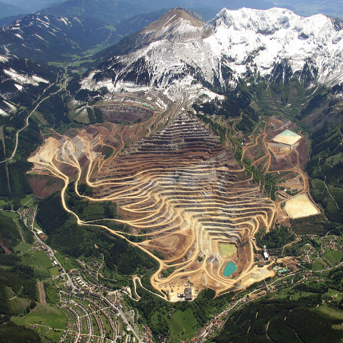 Aerial view of a terraced open-pit mine in a mountain valley, with snow-capped peaks and winding roads around a village.