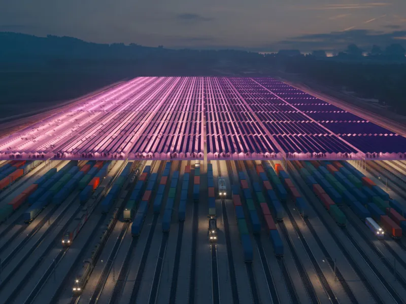 Container yard at dusk with rows of shipping containers and a pink-lit solar-panel canopy over the rail tracks.