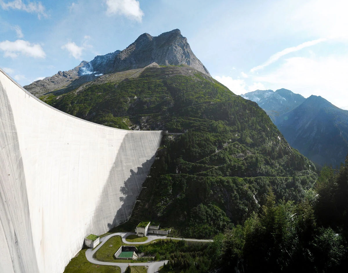 Curved concrete dam spans a forested alpine valley beneath rugged mountain peaks.