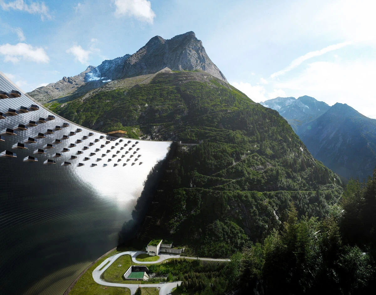 Curved concrete dam along a green mountainside, with a roadway and small buildings at the base, set against rugged alpine peaks and a blue sky.