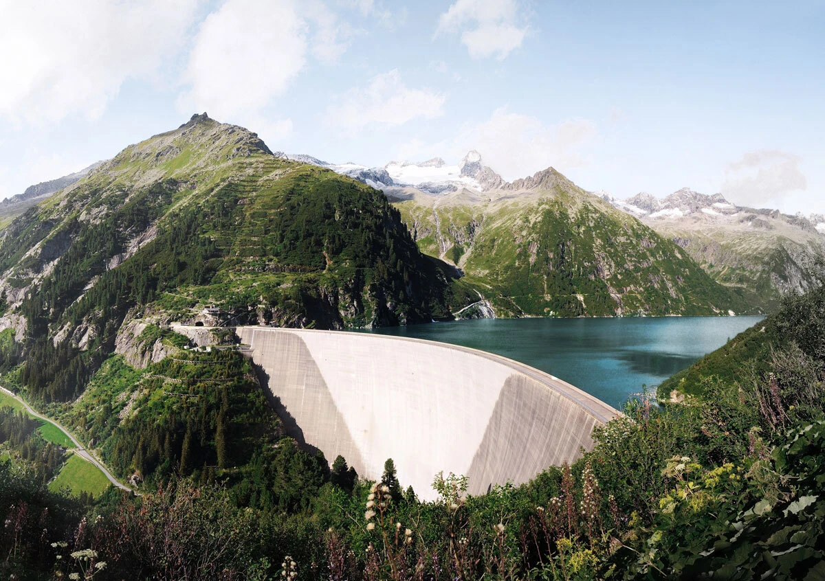 Curved concrete dam spans a turquoise alpine lake in a valley of green, snow-capped mountains.