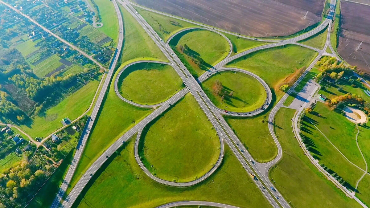 Aerial view of a cloverleaf highway interchange with looping ramps and moving vehicles, surrounded by green fields.