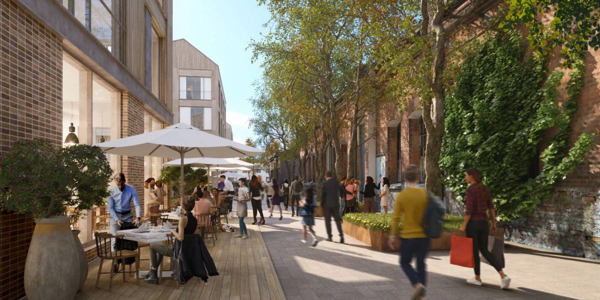 Outdoor cafe scene on a tree-lined city street; people dine at tables under umbrellas while pedestrians stroll by brick buildings.