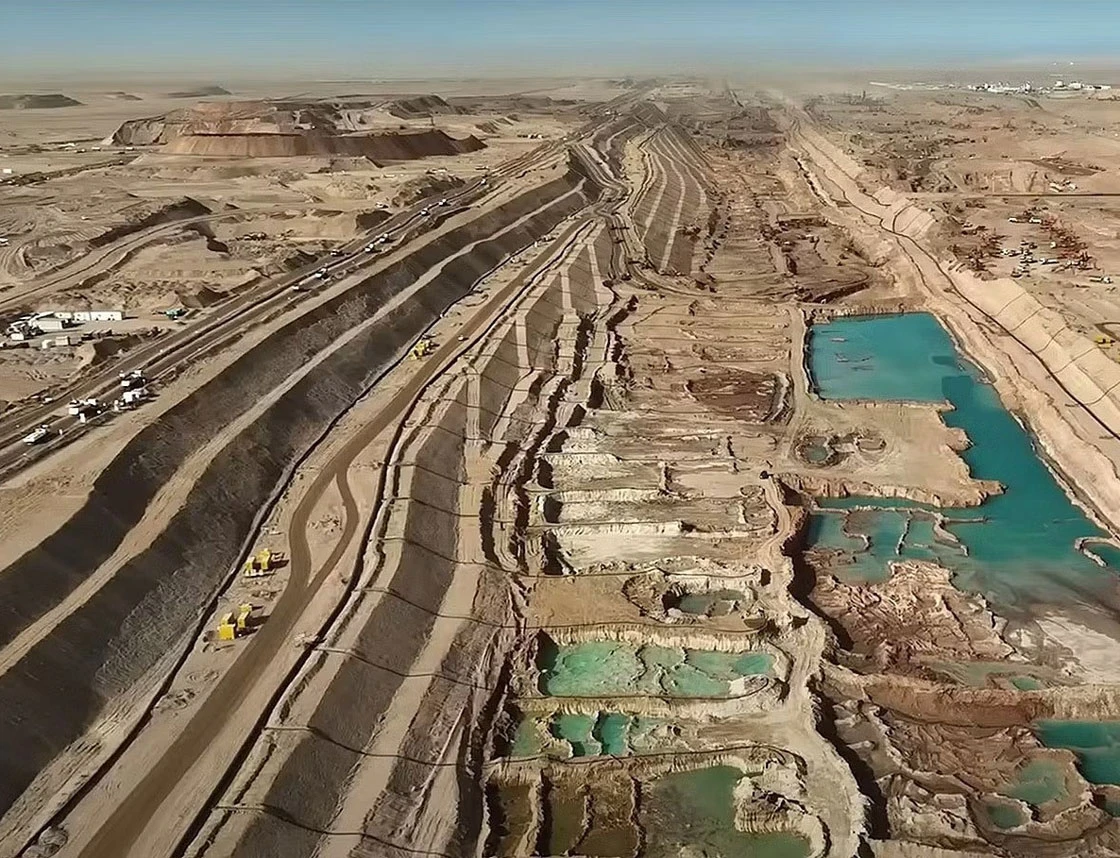 Open-pit mining site in a desert with terraced benches, active earthmoving equipment, and turquoise tailings ponds to the right.