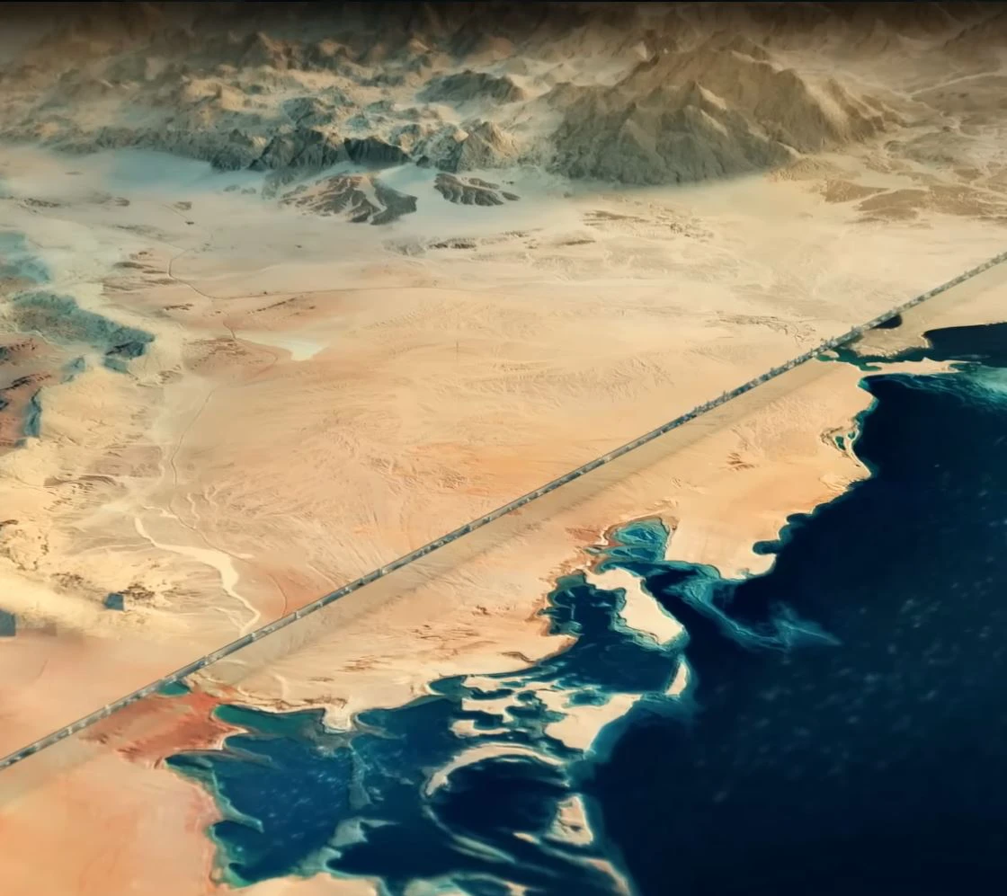 Aerial view of arid desert with a long straight road crossing toward a blue coastline; distant mountains in the background.