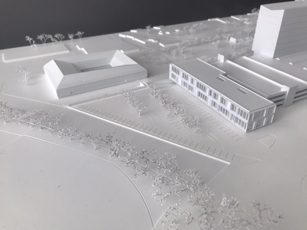 White architectural model of a campus: a glass-fronted rectangular building, a U-shaped block, a parking lot, and surrounding trees.