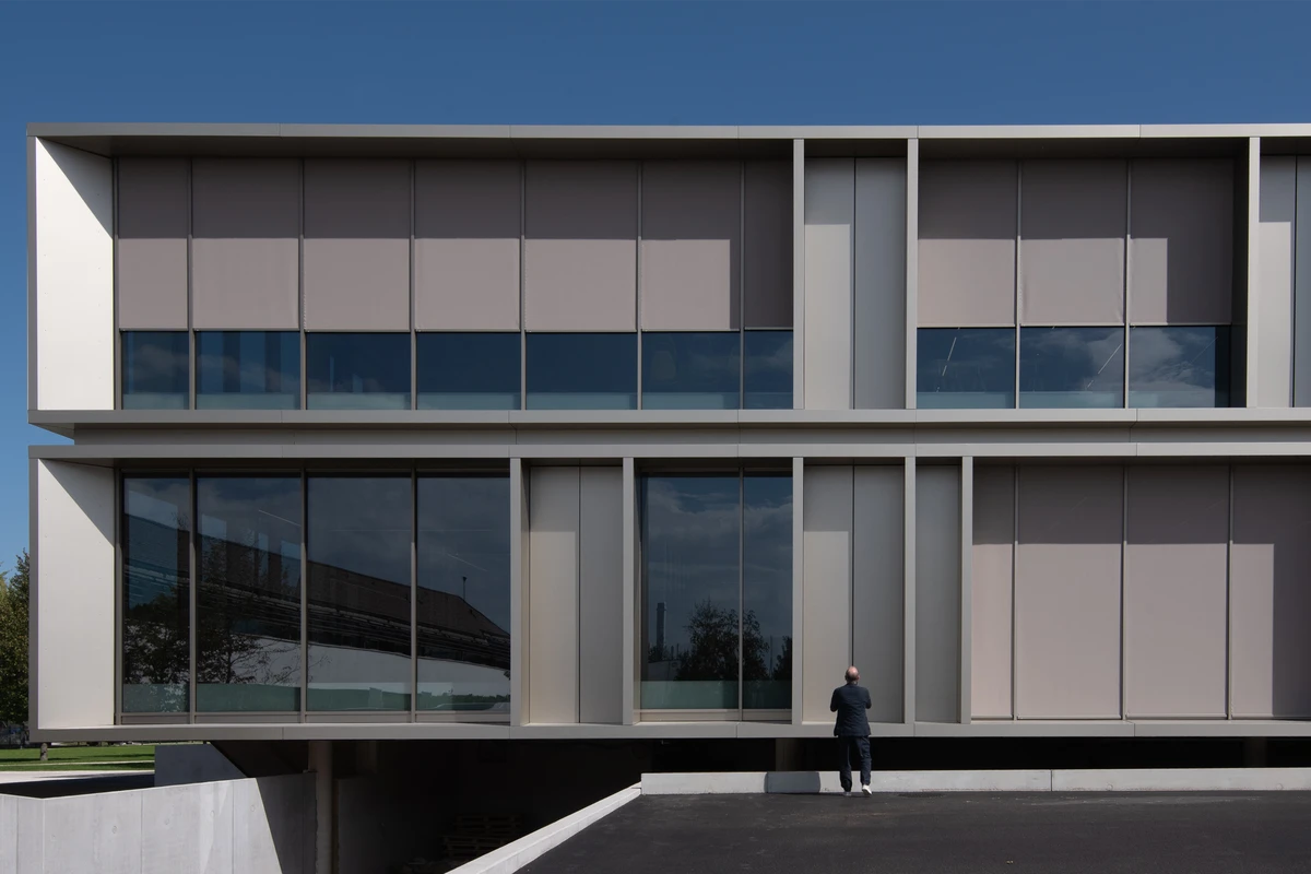 Modern glass-fronted office building with beige panels; a person stands near the entrance, looking at the facade.