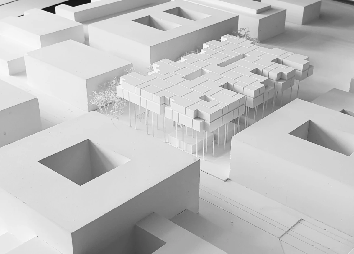 White architectural model of a cantilevered modular cube complex raised on slender stilts, set among blocky buildings with sparse trees.