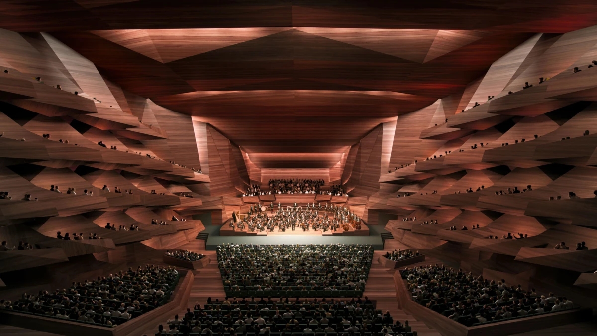 Symphony orchestra performing on a lit stage in a vast wood-paneled concert hall with tiered balconies and audience.