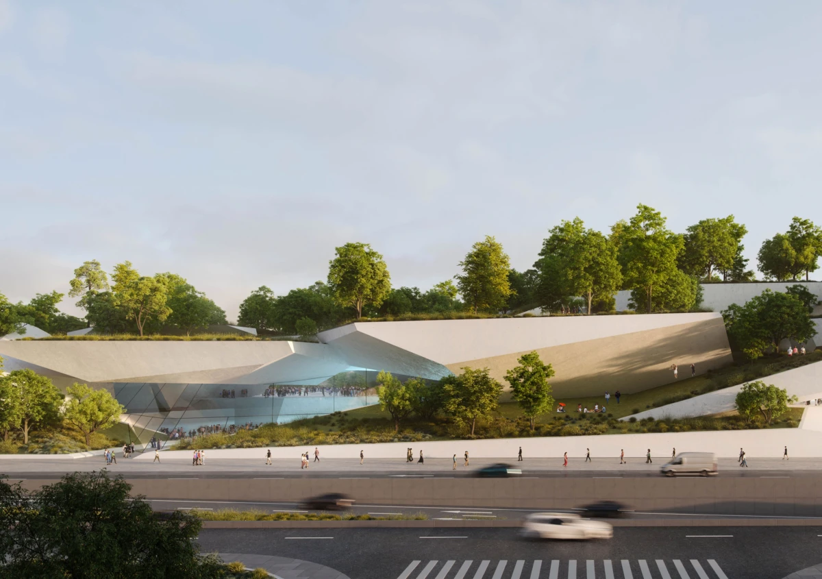 Modern cultural center with angular glass entrance built into a sloped grassy hill; pedestrians on terraces, cars on the highway in daylight.