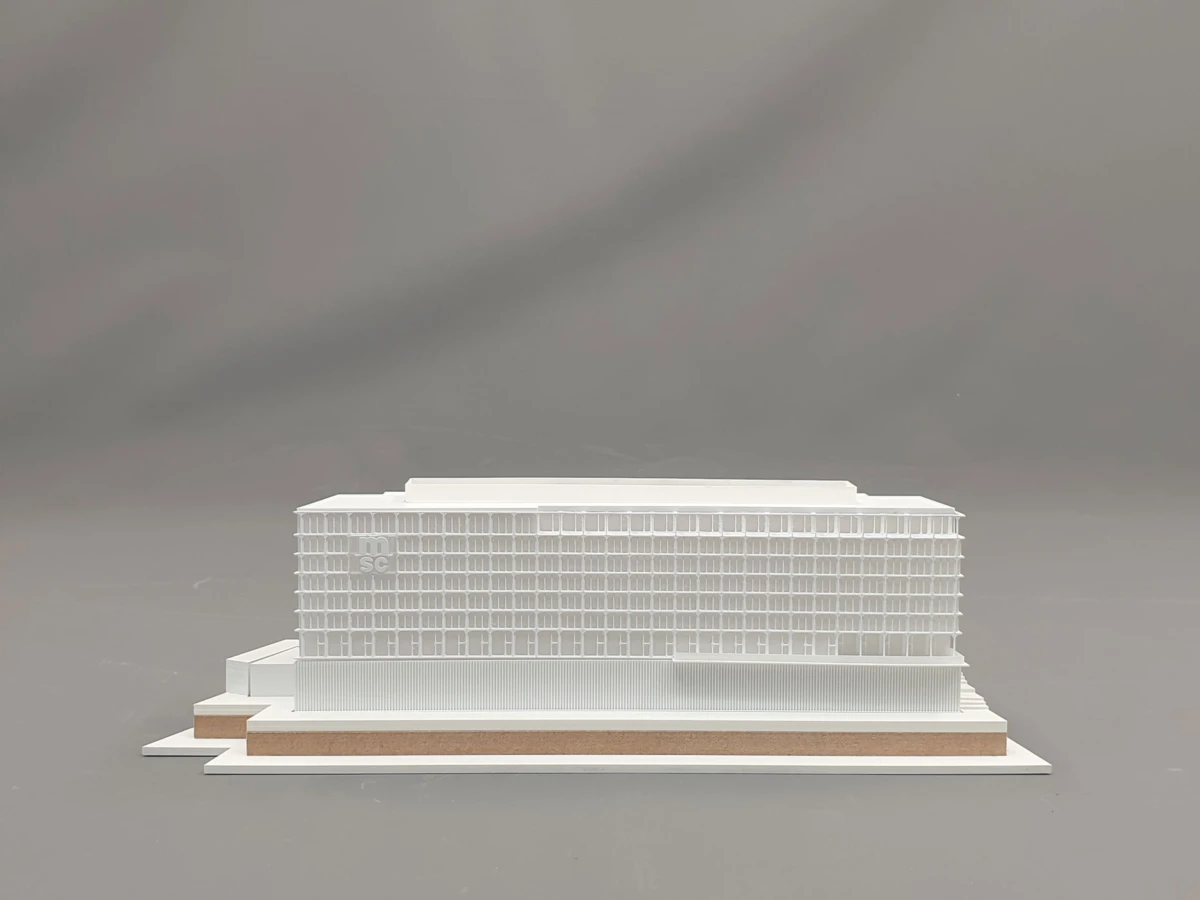 White architectural model of a large multi-story rectangular building with a grid-window facade on a stepped brown and white podium.
