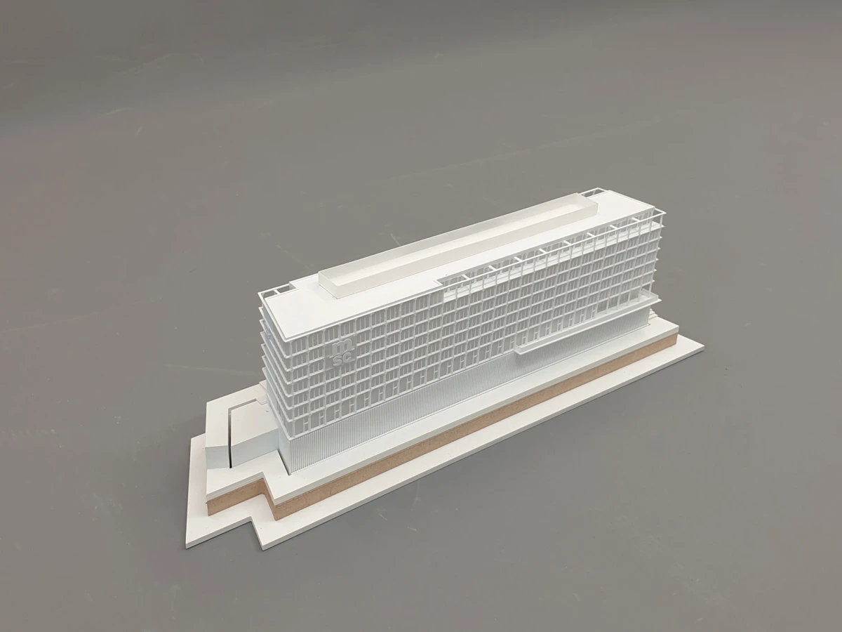 Scale model of a modern, multi-story building with a grid of windows on a stepped base, placed on a gray studio floor.
