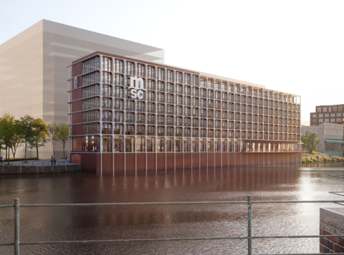 Glass-and-brick office building on a riverfront, with a tree-lined promenade and distant urban blocks.