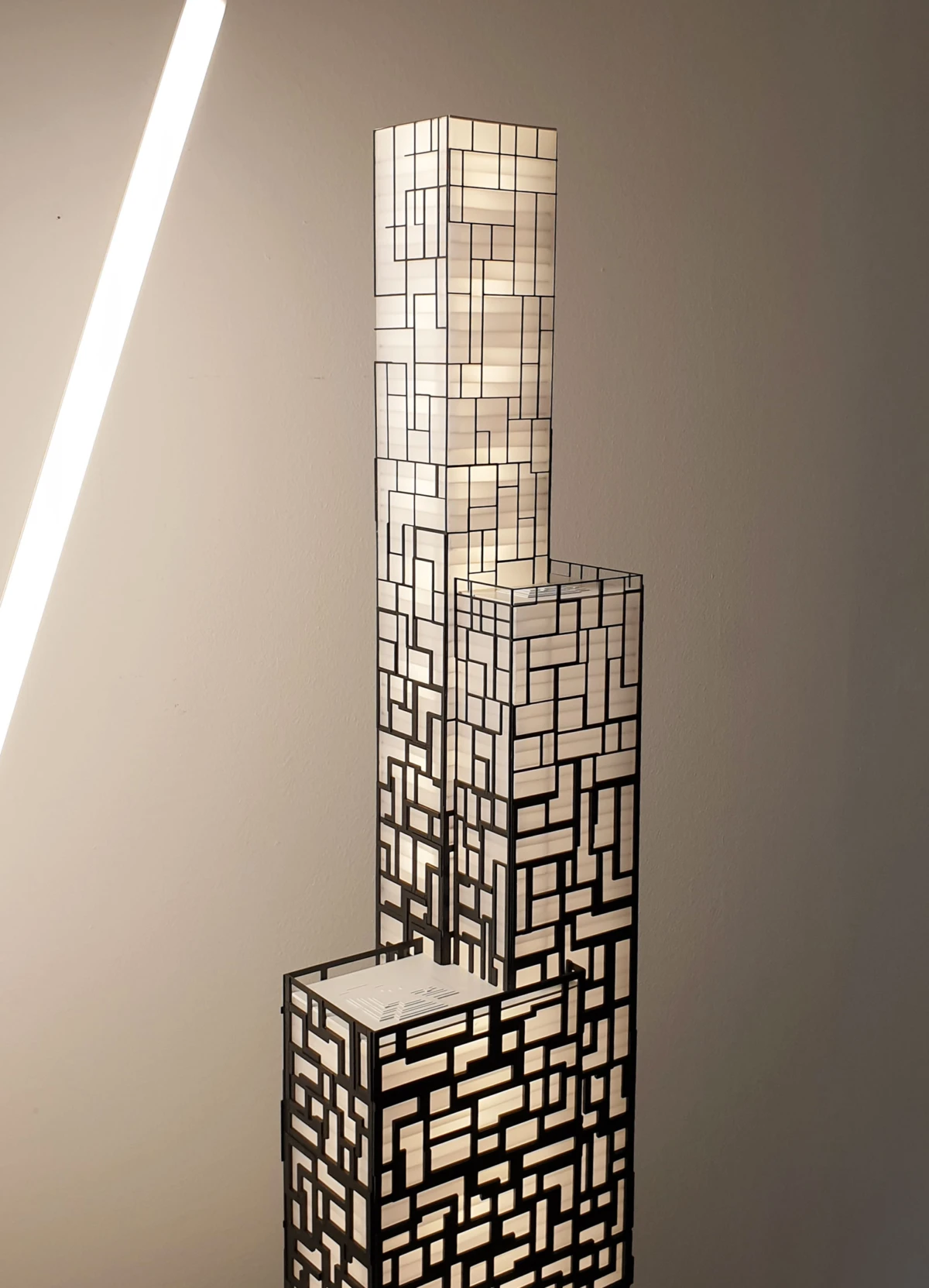 Tall stacked rectangular-lattice sculpture lamp with internal illumination, placed indoors against a plain wall.