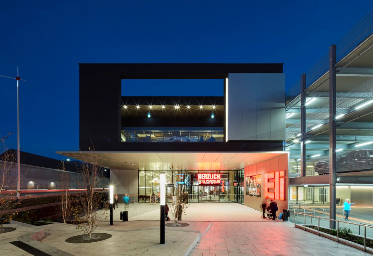 Modern glass-fronted building exterior at dusk with a large black rectangular frame and an illuminated entrance reading HERZLICH WILLKOMMEN.
