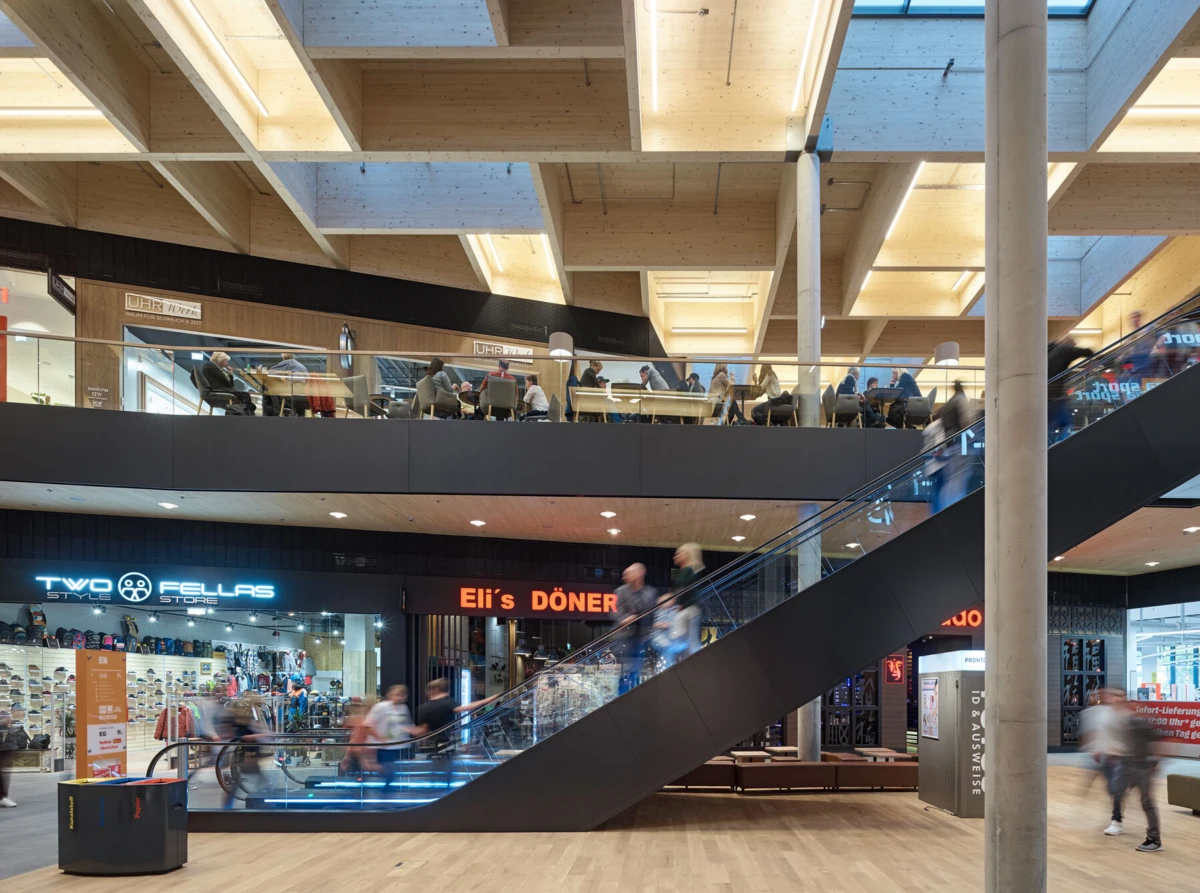 Interior of a modern two-level shopping mall with an escalator; upper-floor seating and storefronts, with pedestrians moving.