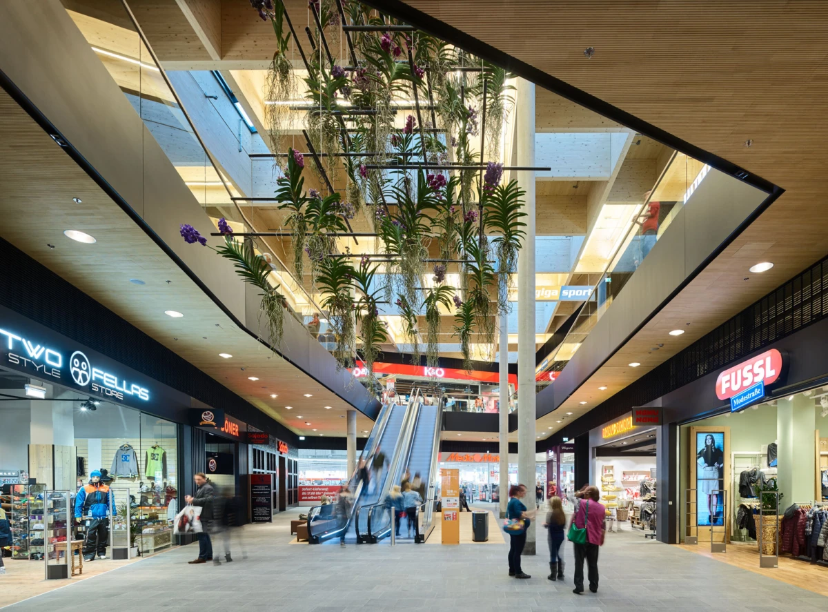 Shoppers walk past storefronts in a modern indoor mall with a central escalator and hanging greenery.