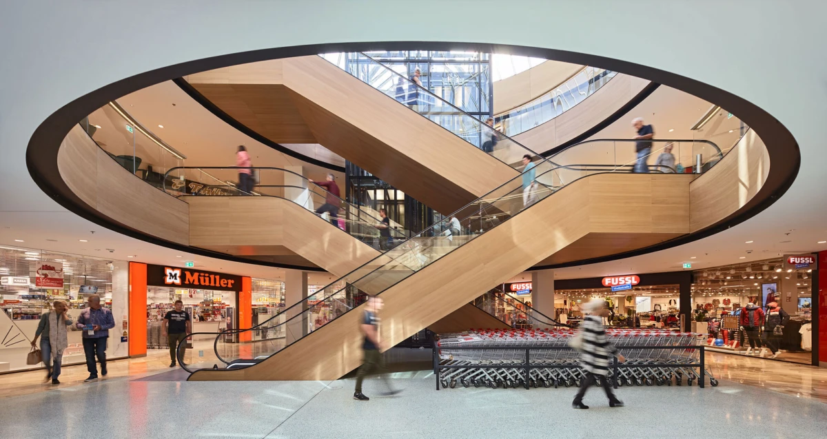 Interior of a modern shopping mall featuring a circular open escalator ring and diagonal escalators, with shoppers on multiple levels.