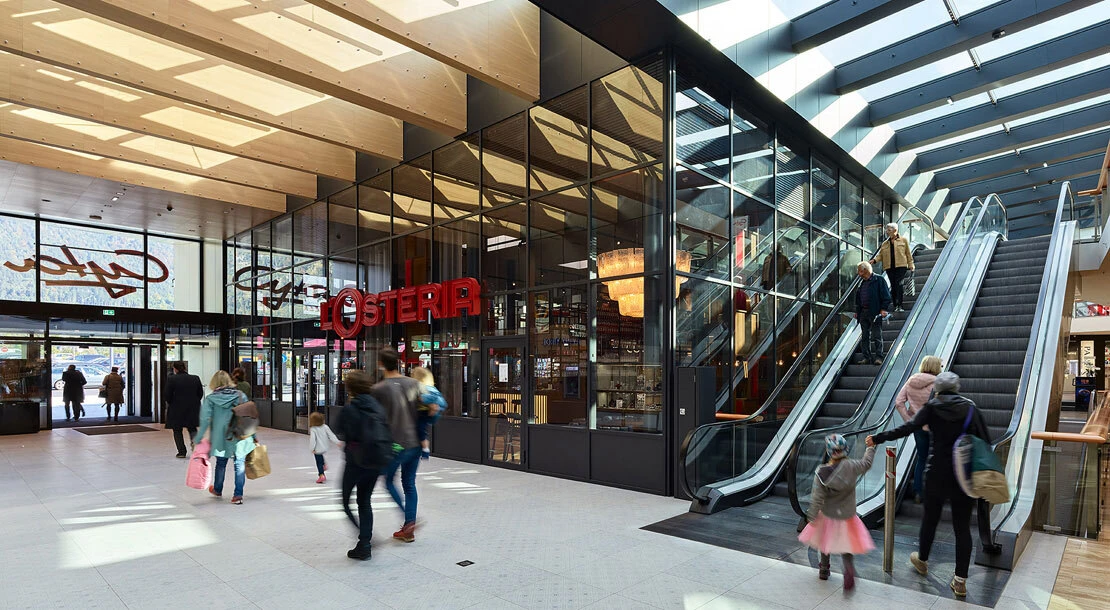 Shoppers walk through a modern shopping mall atrium with glass storefronts and escalators; a red OSTERIA sign visible.