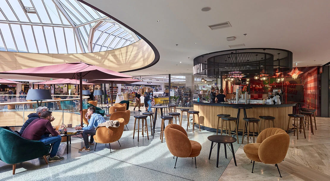 People seated at a circular-bar café in a glass-roofed shopping mall atrium, chatting beneath pink umbrellas.