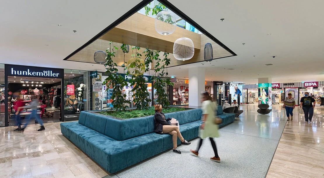 Teal sectional bench surrounds a planter with vines in a mall atrium; a person sits while shoppers pass by under hanging lamps.