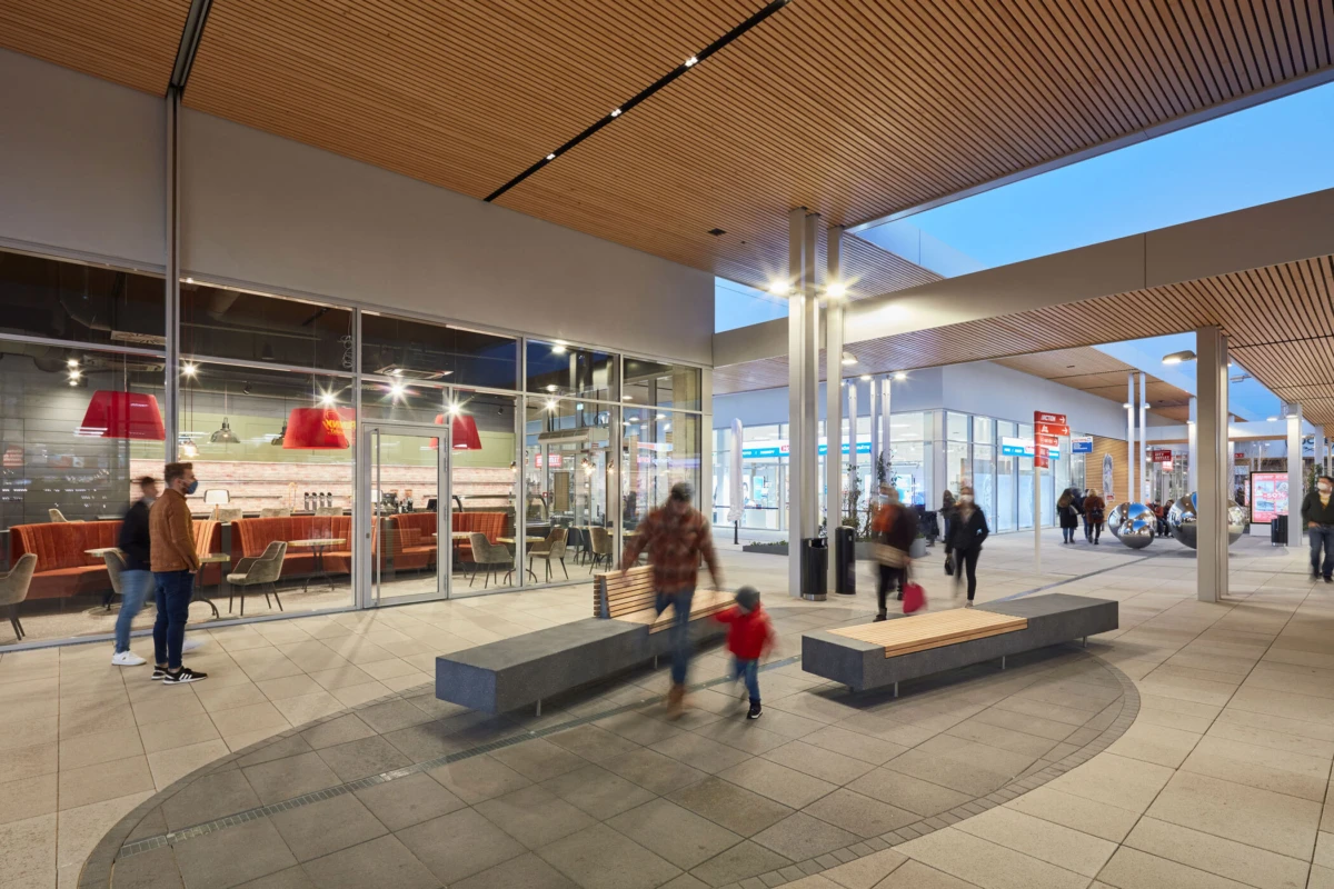Shoppers walk through an indoor shopping mall atrium with glass storefronts, wooden benches, and a cafe visible inside.