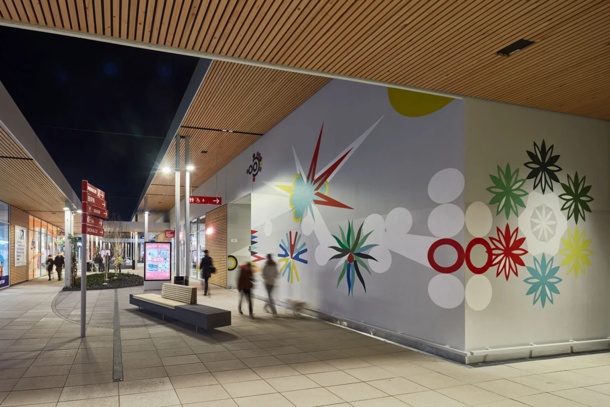 Indoor shopping concourse with a large colorful mural on the right wall; pedestrians walk past benches and signposts.