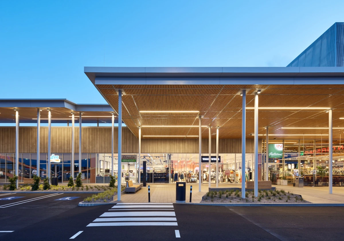 Front view of a modern shopping center entrance with glass storefronts, wooden canopies, and a striped crosswalk in the foreground.