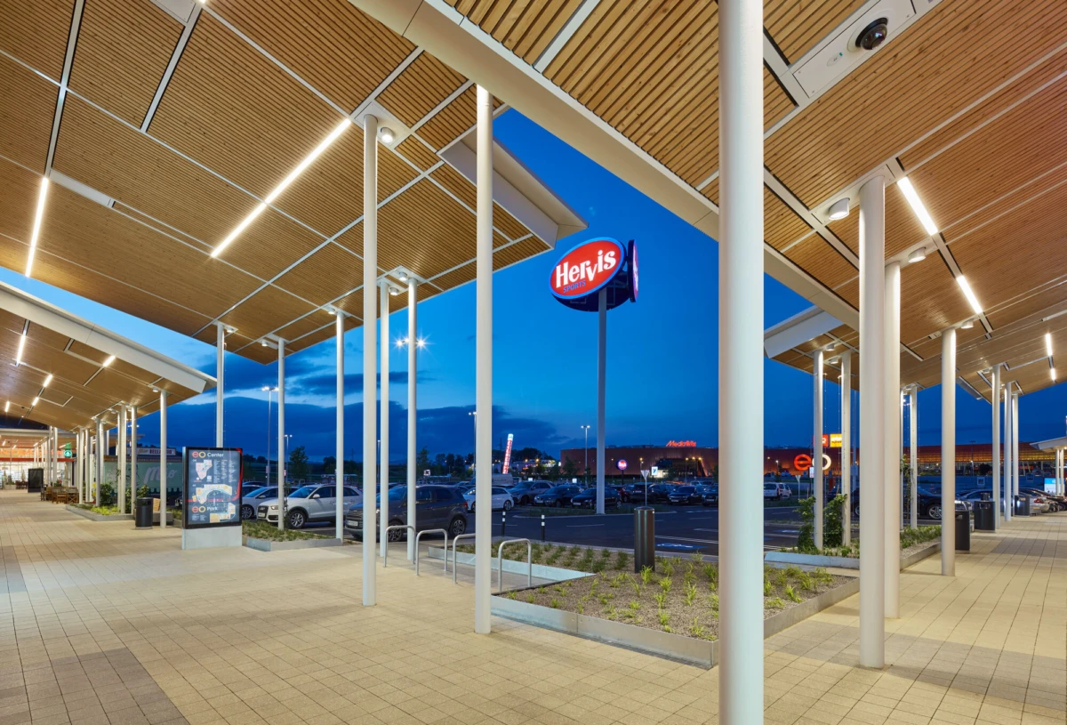 Outdoor shopping center entrance with a covered walkway supported by white columns, empty promenade, and a parking lot at dusk.