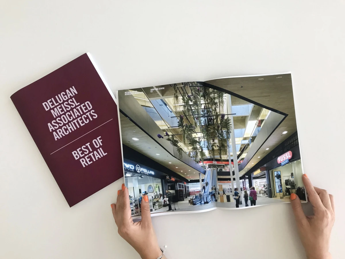 Open architectural magazine showing a shopping mall interior with escalator, hanging plants, and storefronts, held by two hands on a white surface