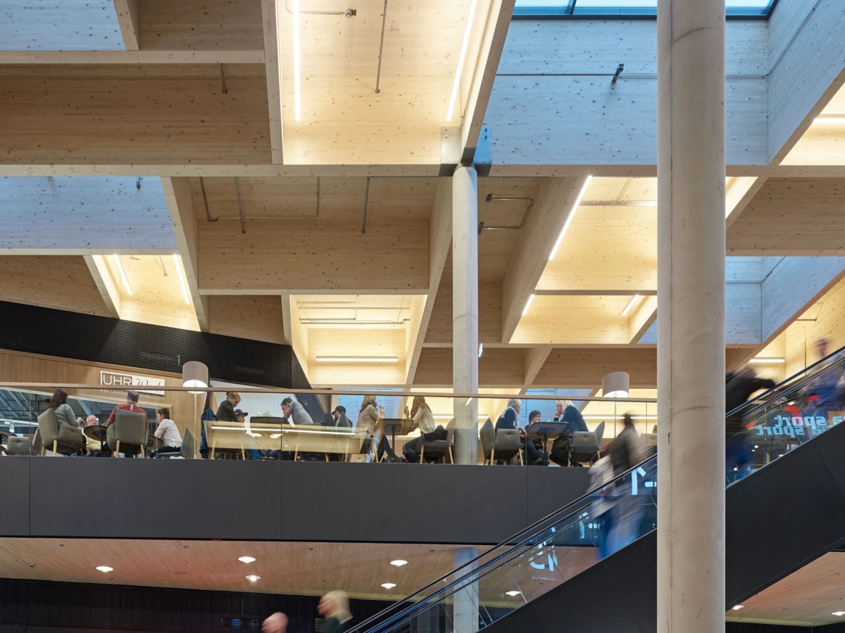 People seated at a long table on a mezzanine in a bright, modern wood-and-concrete interior with skylights and an escalator.