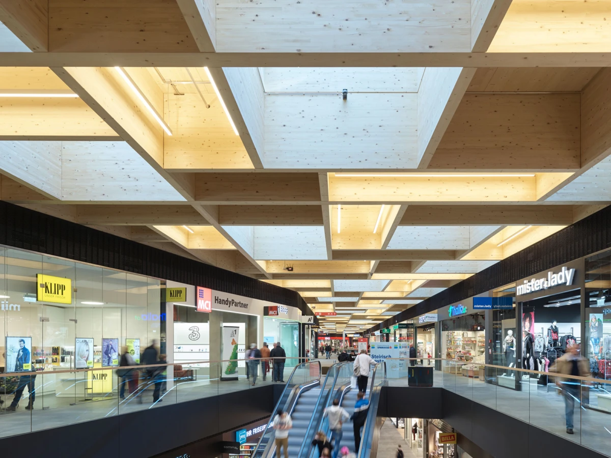 Two-level shopping mall interior with escalators, glass railings, and a wooden grid ceiling; shoppers walk past storefronts.