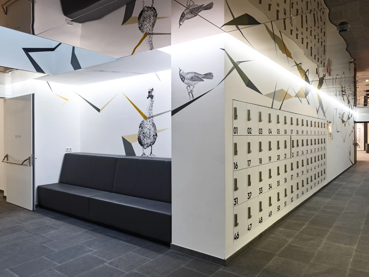 Long hallway with numbered lockers along a white wall, geometric mural with birds; black leather sofa on the left.