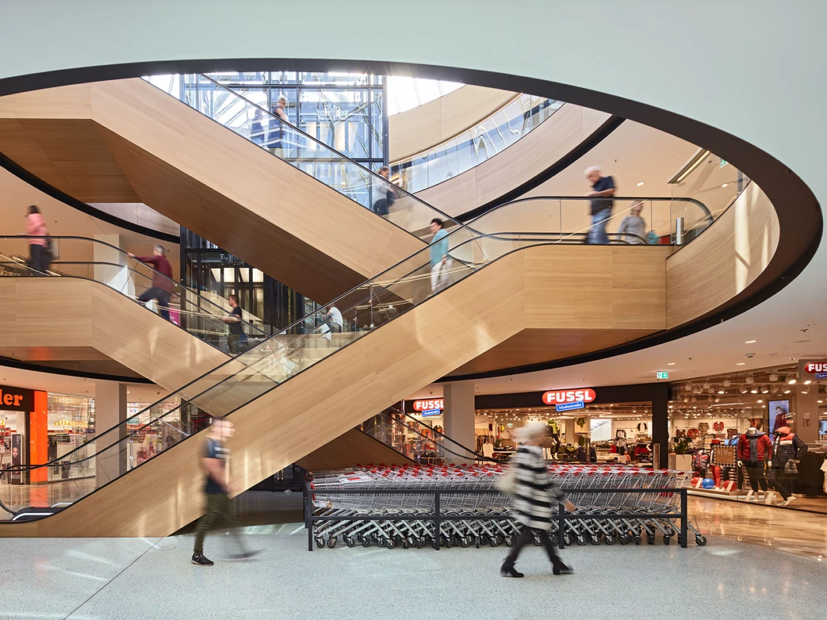 Wooden looping escalators form a circular atrium in a shopping mall, with shoppers riding and walking on each level.