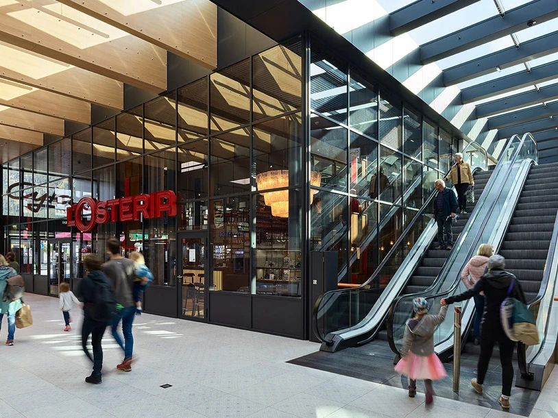 Shoppers walk through a modern shopping mall beside a glass-front store and an escalator.