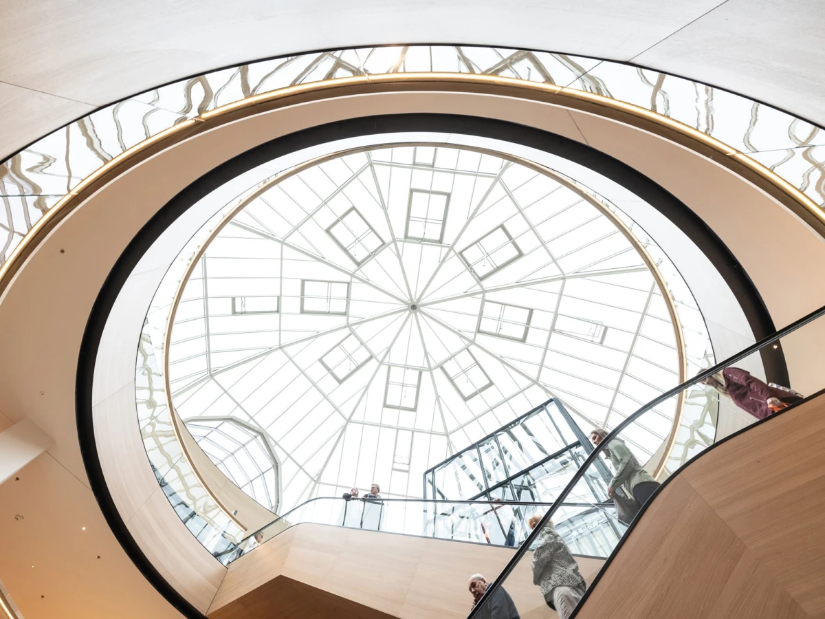 People stand along the glass-railed spiral staircase in a circular glass-domed atrium with a geometric skylight overhead
