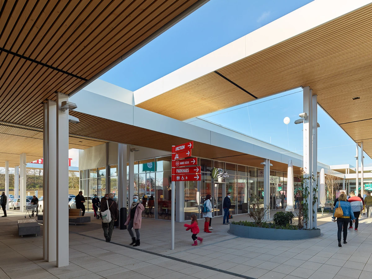 Shoppers walk through a modern outdoor shopping plaza under a wooden-slat canopy; storefronts and red directional signs visible.