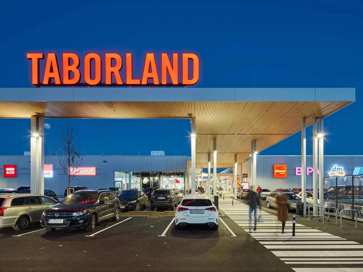 TABORLAND storefront with large orange sign, covered entrance, pedestrians crossing at a zebra-crosswalk in a parking lot at dusk.
