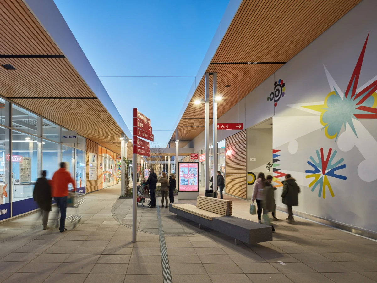 Shoppers walk along a modern shopping mall corridor lined with storefronts and a colorful mural on the right.